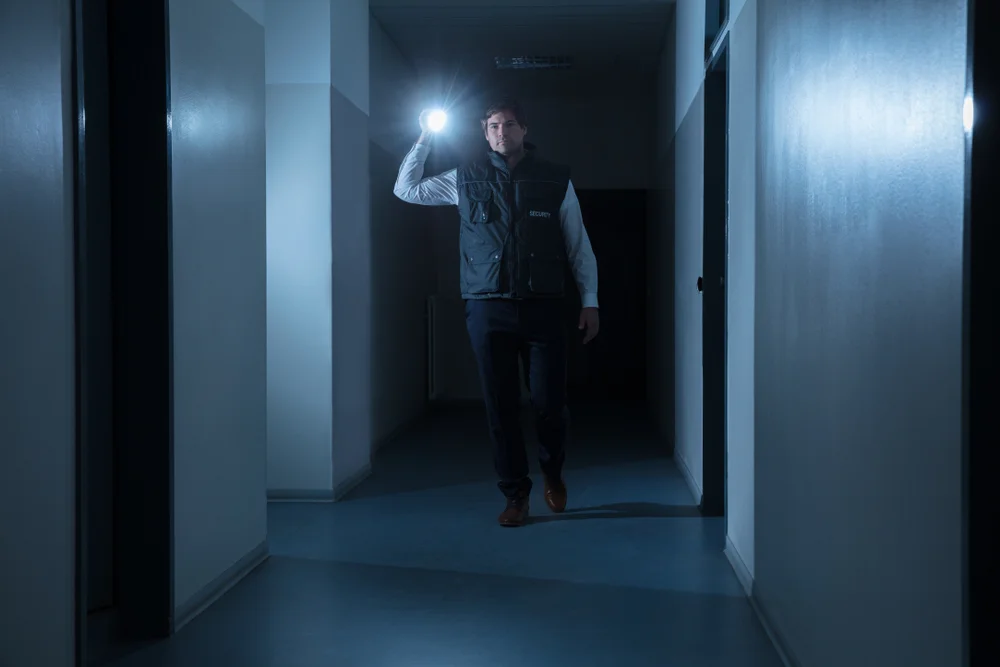 A security guard walking through a dark hallway with his flashlight in his hands.