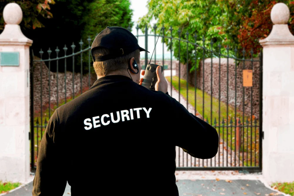 A security guard standing in front of the gate of a house while talking on the radio.