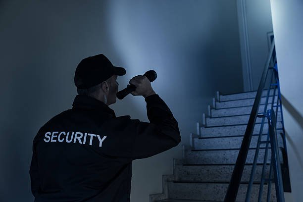 A man with a security jacket looking towards a flight of stairs with his flashlight.
