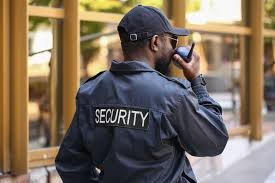 A security guard facing away from the camera and holding a radio close to his face to respond.